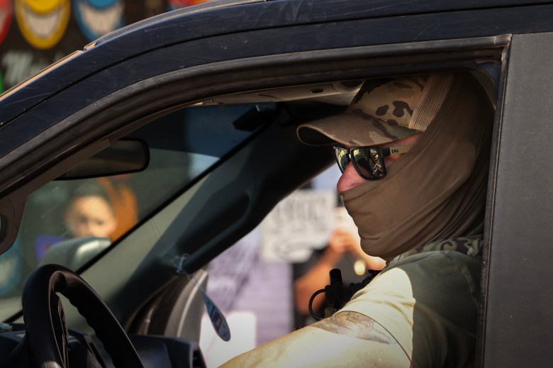A person wearing military-style camouflage, sunglasses, and a tan balaclava sits in the driver's seat of a dark vehicle, facing forward. Their arm rests on the open window. The reflection in their sunglasses reveals figures in the street. A child's face is visible through the passenger window, slightly out of focus. The background features colorful cartoonish smiley faces painted on a wall.